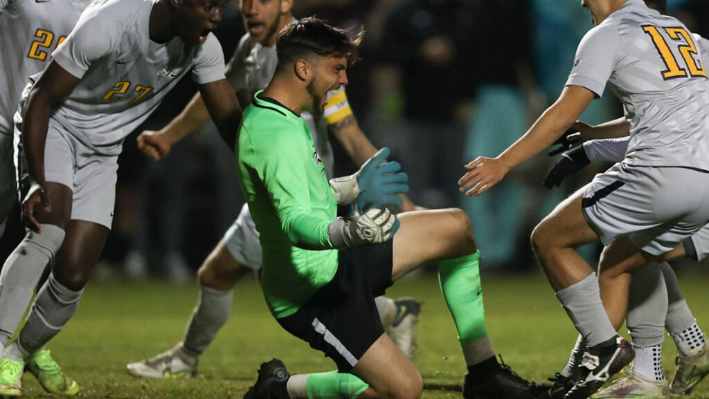 UNCG men's soccer player celebrates on the field, surrounded by Stanford players.